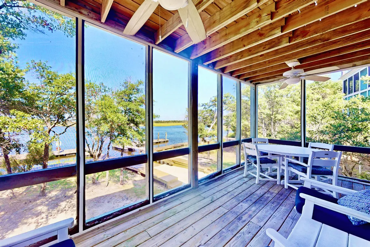 The image shows a serene interior living space with a wooden ceiling, large windows that frame sweeping water views, and white outdoor furniture arranged for relaxation. Soft morning light filters in, highlighting the inviting setting that's perfect for coffee at sunrise or evening gatherings with friends. The space feels open, airy, and seamlessly connected to the marsh beyond.