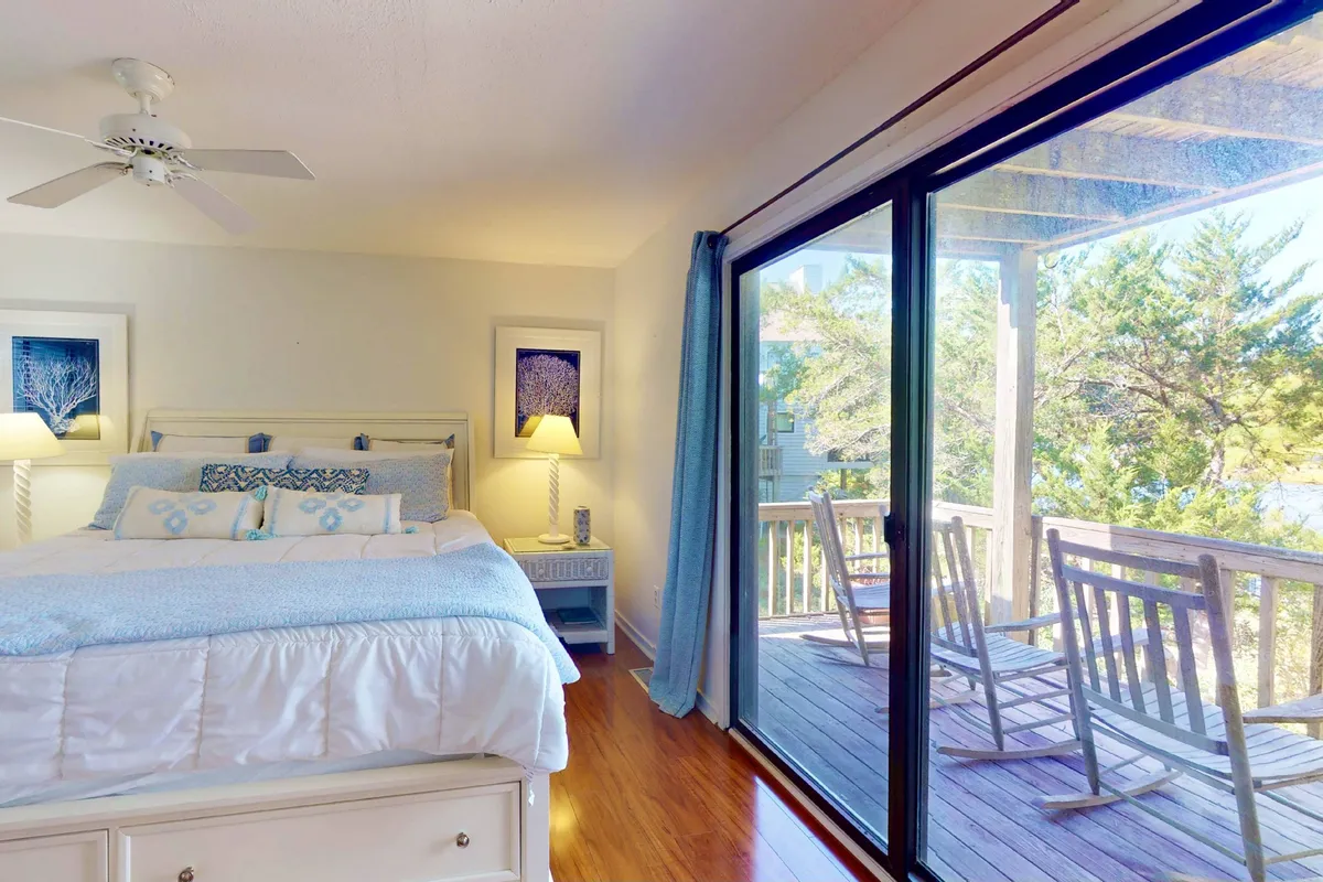 The primary bedroom opens onto a deck with rocking chairs, seen through large sliding glass doors. The bed is adorned with decorative pillows, and the space is bathed in sunlight, with hints of nature visible just outside.