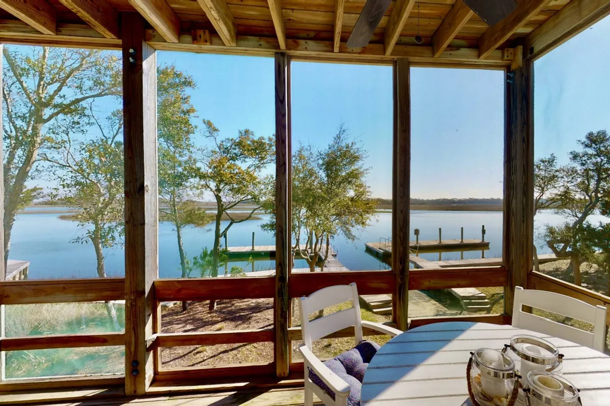 A serene screened porch with a dining table overlooking tranquil water and lush trees. The scene is peaceful, with soft natural light, inviting guests to sit and relax while enjoying the outdoors without exposure to the elements.