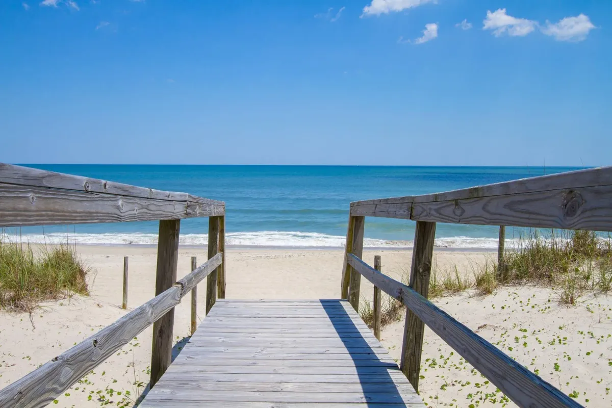 A wooden walkway extends over the dunes, leading directly to the sandy beach and the expansive ocean beyond. The sky is clear, and the horizon is visible in the distance, emphasizing the tranquil seaside environment.