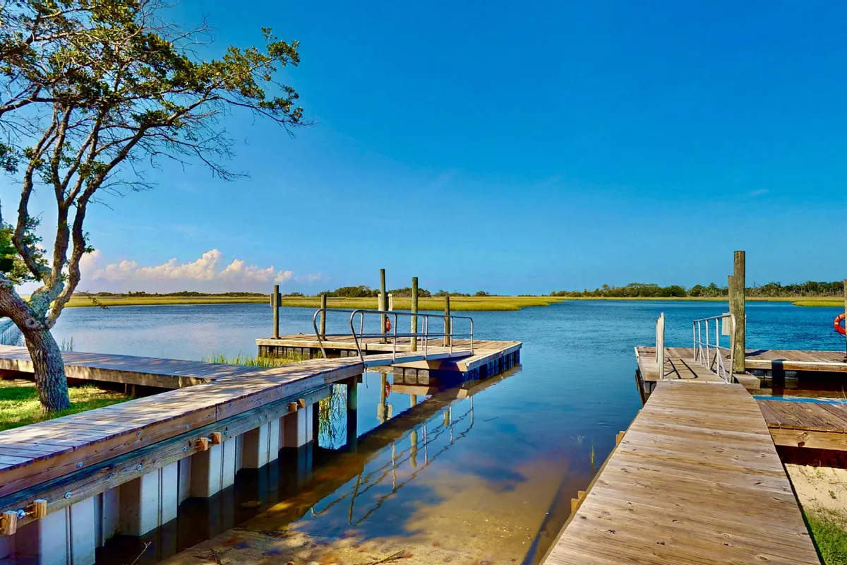 A peaceful scene featuring docks stretching out over calm water, a tree providing shade, and a backdrop of blue sky. The tranquil environment invites visitors to connect with nature, fish, or simply enjoy the view.