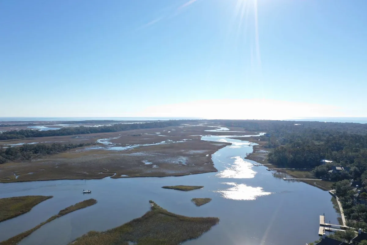 An expansive view of winding waterways snaking through lush marshland, with a small boat navigating the tranquil waters and a distant tree line. The scene evokes the serenity and wild beauty of the salt marsh, ideal for boating, bird watching, and soaking in nature.