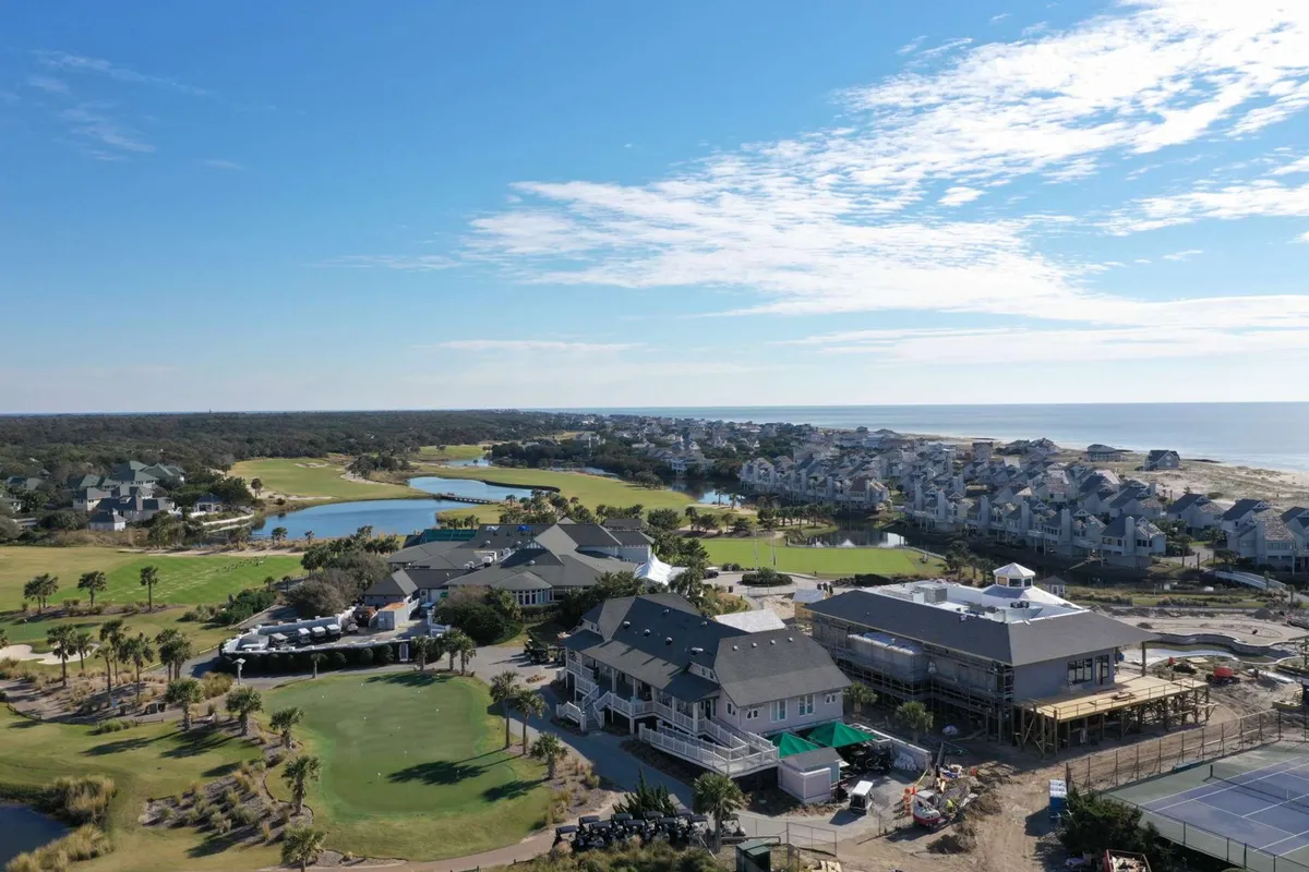 A broad, panoramic view of the island’s golf course, clubhouse, nearby beach, and residential area, framed by water features. The scene conveys the breadth of recreational amenities available on Bald Head Island.