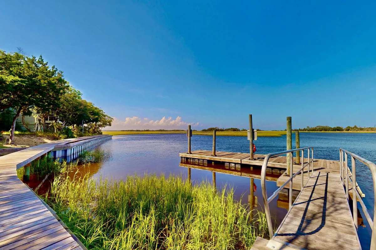 A serene waterfront dock bordered by a wooden boardwalk and surrounded by lush greenery, stretching out over calm waters under an open sky. The dock invites activities like fishing, launching kayaks, or simply enjoying the peaceful marsh setting.