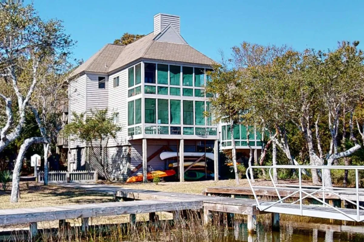 A multi-level house overlooks a dock, with colorful kayaks ready for use and expansive marsh views. The water glows with sunrise or sunset light, and the scene exudes peaceful outdoor adventure, including kayaking and fishing.