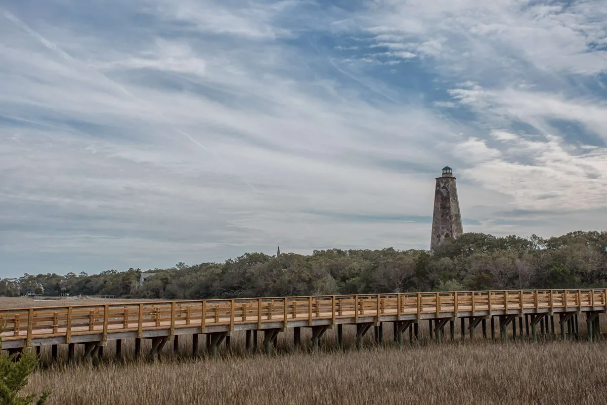 This image captures a scenic view of the iconic Bald Head Island lighthouse, a wooden boardwalk, and surrounding marshland. The tranquil setting emphasizes the island’s unique landscape and opportunities for walking, wildlife observation, and photography.