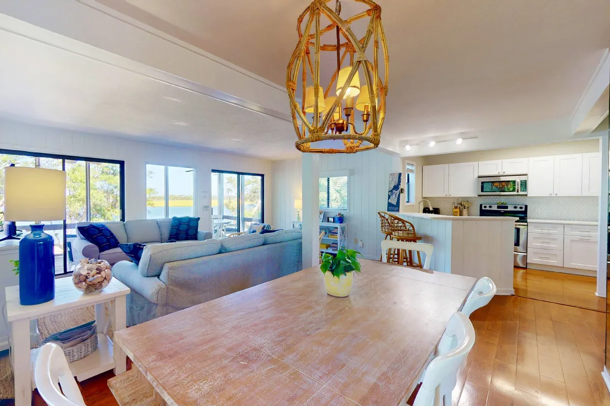 An open-concept dining and living area with a dining table, sofa, chandelier, and kitchen in view. Large windows bring in natural light, creating a warm, communal space ideal for meals, conversation, and planning outings.