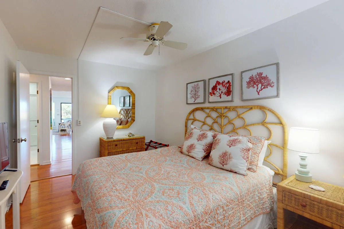 A serene queen bedroom decorated with coral-patterned bedding, tasteful wall art, a rattan headboard, a table lamp, and a wooden dresser. Sunlight fills the room, evoking a calm and welcoming ambiance for restful nights.