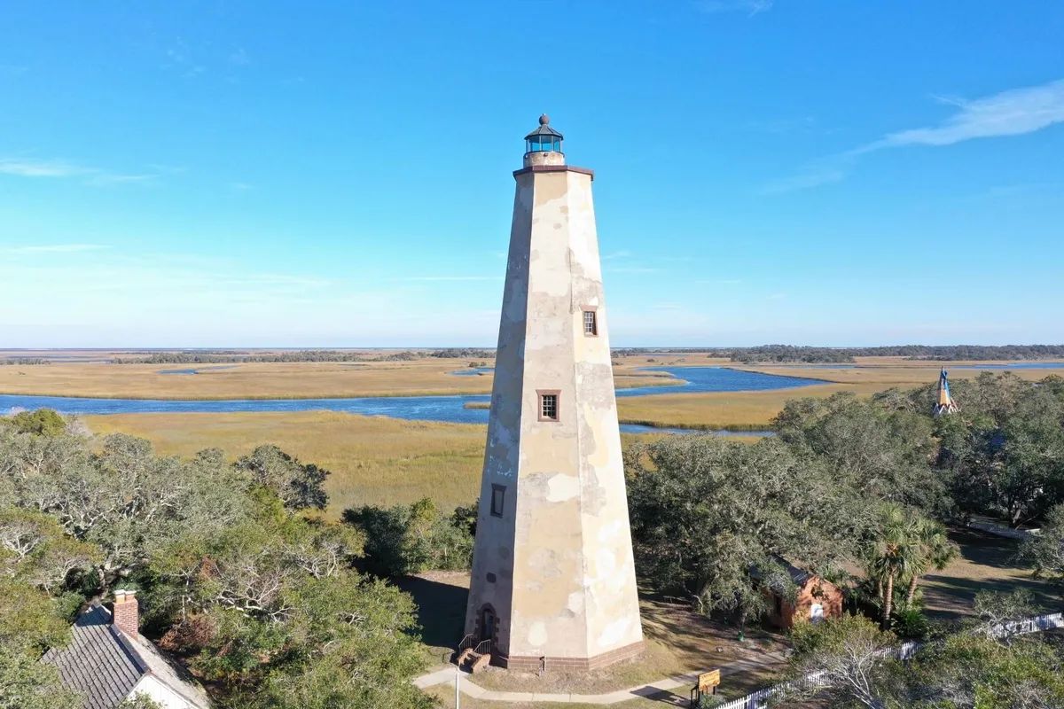 The iconic Old Baldy Lighthouse rises above surrounding marshland under a brilliant blue sky. The area is open and natural, inviting exploration, tours, and opportunities for memorable photographs.